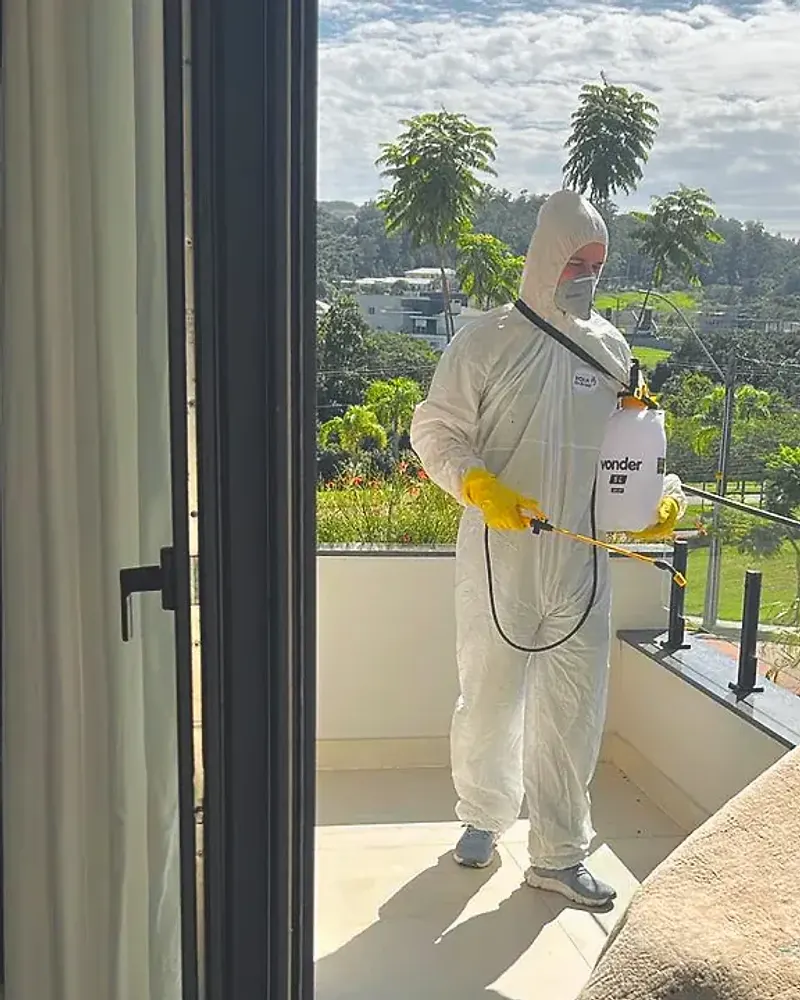 Técnico de macacão branco e pulverizador costal aplica produto na varanda de um apartamento, visto de dentro através da porta de correr aberta.