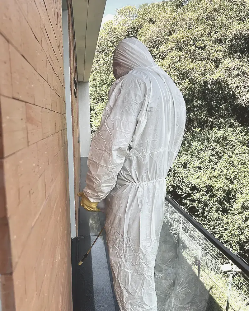 Técnico de macacão branco aplica produto ao longo do rodapé externo de uma sacada, com mata ao fundo.